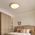 A cozy bedroom with a large bed, earth-toned pillows and blanket, wooden headboard, built-in wardrobe, and wall shelves. Natural travertine accents and the Wabi-Sabi Travertine LED Ceiling Light create a minimalist warmth.