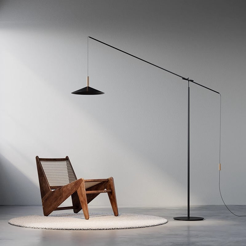 A minimalist room features an angular wooden chair with a woven seat. The sleek black Velluto Floor Lamp arches overhead, providing adjustable lighting on the round beige rug, while walls and floor exhibit neutral tones.