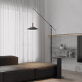 A minimalist living room showcases a dark brown sectional sofa and the sleek Velluto Floor Lamp. Sheer white curtains and a modern side table add charm, while light gray walls with built-in shelves and adjustable lighting enhance the contemporary design.