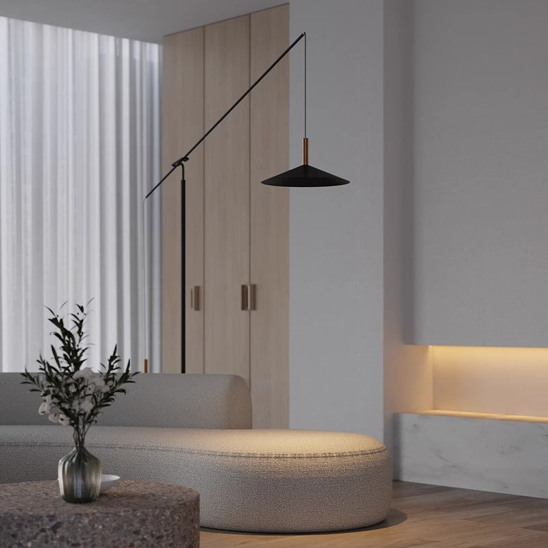 A modern living room with minimalist decor features a beige curved sofa, the black Velluto Floor Lamp, wooden paneling, adjustable lighting, and a stone table holding a round vase with greenery. White curtains adorn the windows.