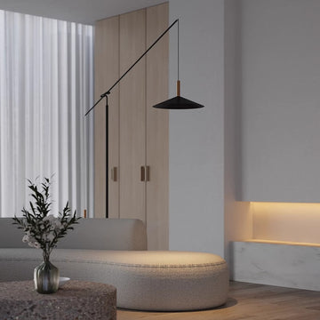 A modern living room with minimalist decor features a beige curved sofa, the black Velluto Floor Lamp, wooden paneling, adjustable lighting, and a stone table holding a round vase with greenery. White curtains adorn the windows.
