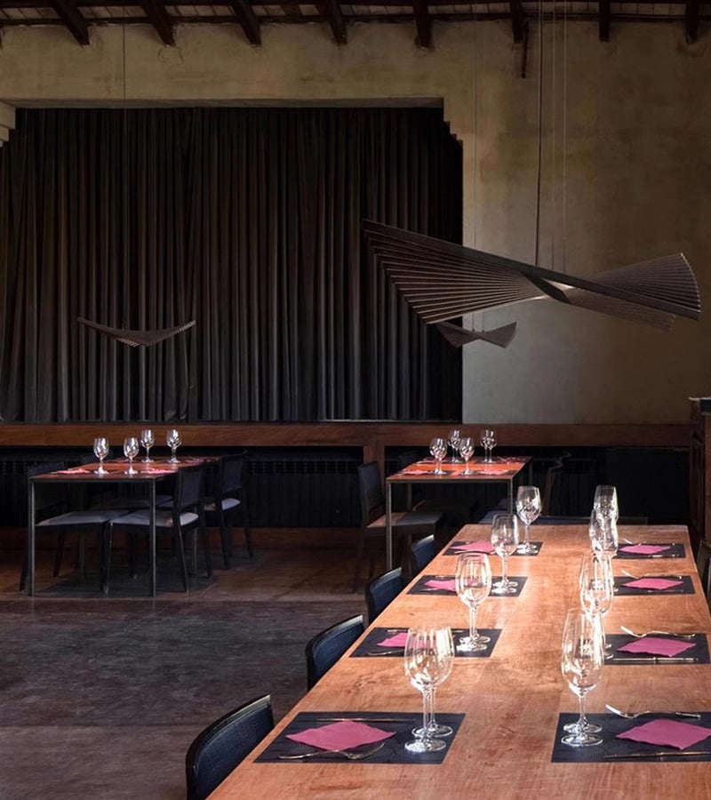 A dimly lit restaurant showcases a long wooden table set with wine glasses, black placemats, and red napkins. The elegant Fishbone Wave Chandelier casts a sophisticated glow against the large, dark curtain covering the back wall.