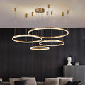 Modern chandelier with multiple circular LED rings hanging from the ceiling in a dark room with a gray sectional sofa and decorative elements on shelves.