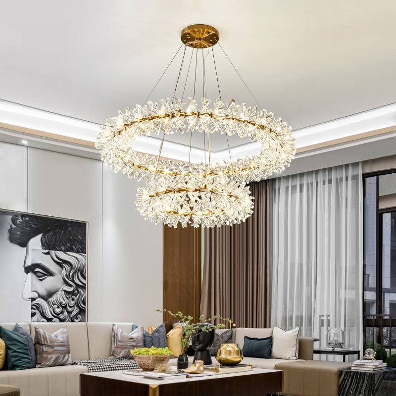 A modern living room showcases a Crystal Branch Chandelier with adjustable wire, golden accents, and crystal details. Below it, a beige sectional sofa with various cushions encircles a wooden coffee table, while a large portrait adorns the wall.
