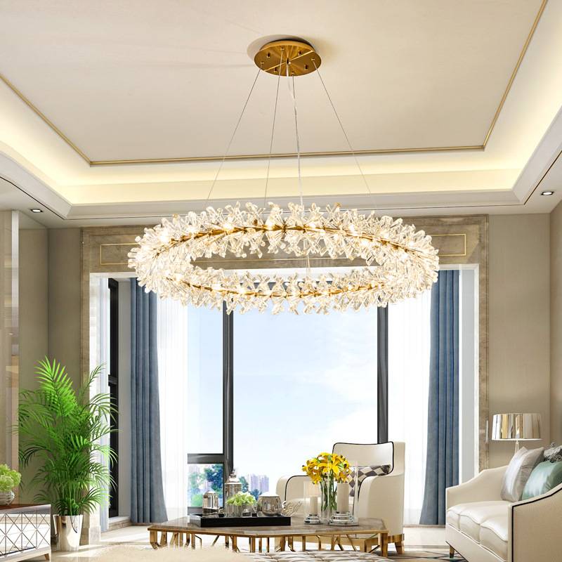 A luxurious living room showcases a Crystal Branch Chandelier with its adjustable wire and energy-efficient G4 LED bulbs, floor-to-ceiling windows adorned with blue curtains, a white armchair, potted plants, and a table with yellow flowers. The decor is dominated by neutral tones.