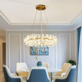 A contemporary dining room features a round white table with five cushioned chairs, enhanced by the Crystal Branch Chandelier. Its adjustable wire complements light gray walls, while energy-efficient LED bulbs illuminate a corner painting and potted plant.