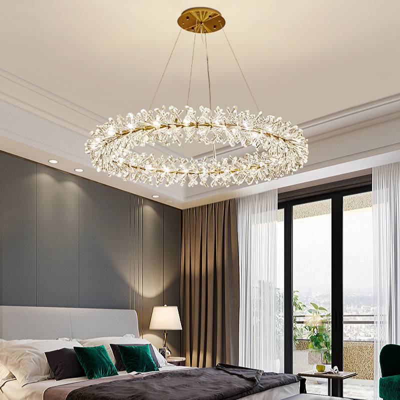 A modern bedroom showcases the large Crystal Branch Chandelier, featuring crystal accents and energy-efficient LED bulbs with adjustable wire. The room also includes a neatly made bed, side table with lamp, and floor-to-ceiling windows adorned with sheer and heavy curtains.