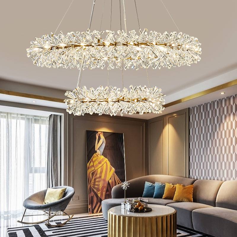 The modern living room dazzles with a Crystal Branch Chandelier and G4 LED bulbs, reflecting elegantly. It boasts a curved gray sofa adorned with multicolored pillows, a striped rug, gold coffee table, and yellow abstract painting. Large windows with sheer curtains allow natural light to flood in.