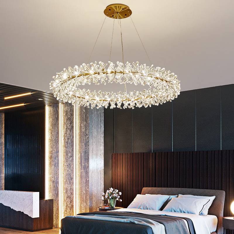 A modern bedroom features a sophisticated Crystal Branch Chandelier with adjustable wire and crystal-like elements, dark wood-panelled wall, gray and white bedding on the bed, and an elegant flower arrangement on the bedside table.