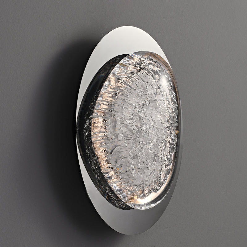 The wall lamps circular design, with textured glass like ice, complements an energy-efficient LED light creating an elegant look. It is ideally suited for bedroom use and mounts seamlessly on a smooth dark gray wall.
