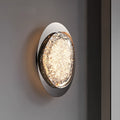 A wall lamps for bedroom features a circular design with a textured, luminous surface, an energy-efficient LED, and a reflective metal frame. Mounted on a neutral wall, it emits a warm glow.