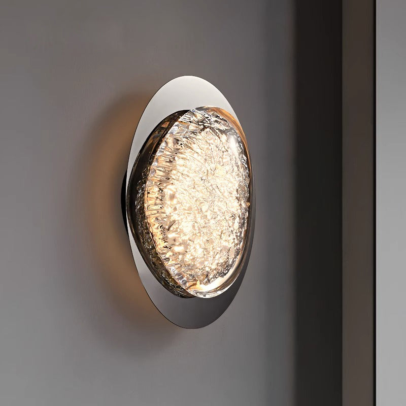 A wall lamps for bedroom features a circular design with a textured, luminous surface, an energy-efficient LED, and a reflective metal frame. Mounted on a neutral wall, it emits a warm glow.