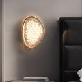 The wall lamps for bedroom offer a unique, modern acrylic design with an irregular oval shape that emits a warm glow. Featuring smart dimming, theyre mounted next to a dark upholstered headboard. Nearby, a small stack of books sits on a table amidst the minimalist decor that enhances their elegance.